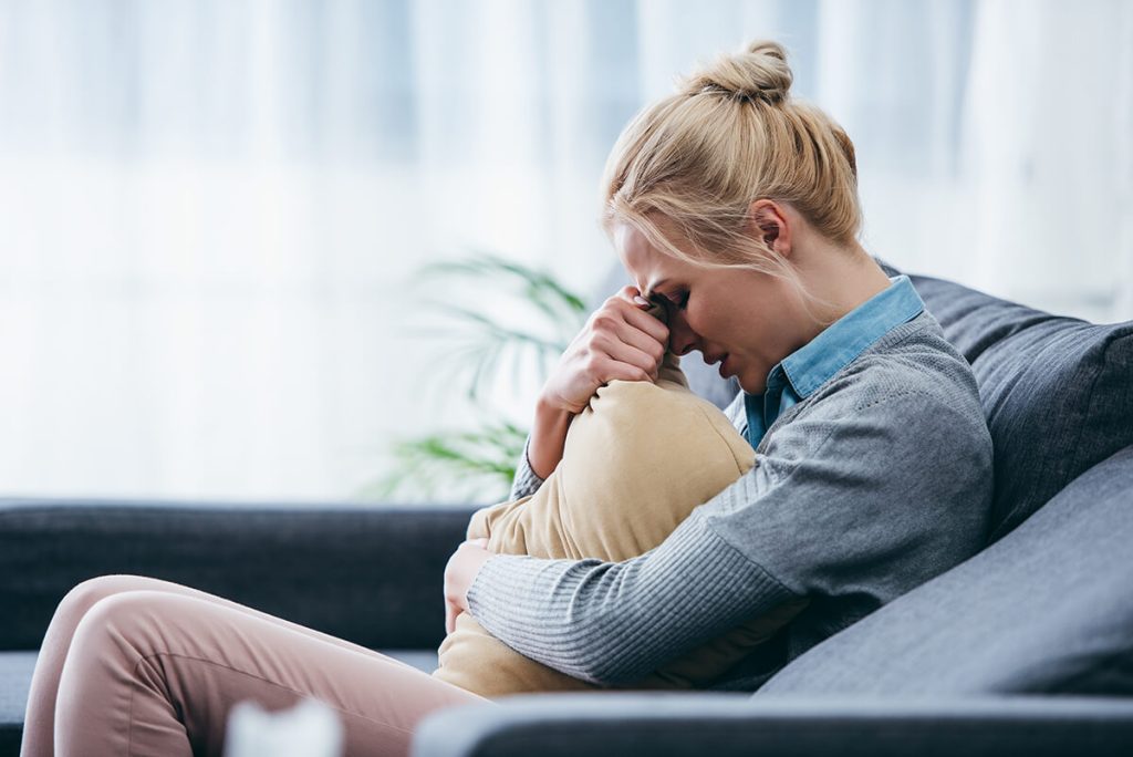 Sad woman sitting on couch, crying and holding pillow at home