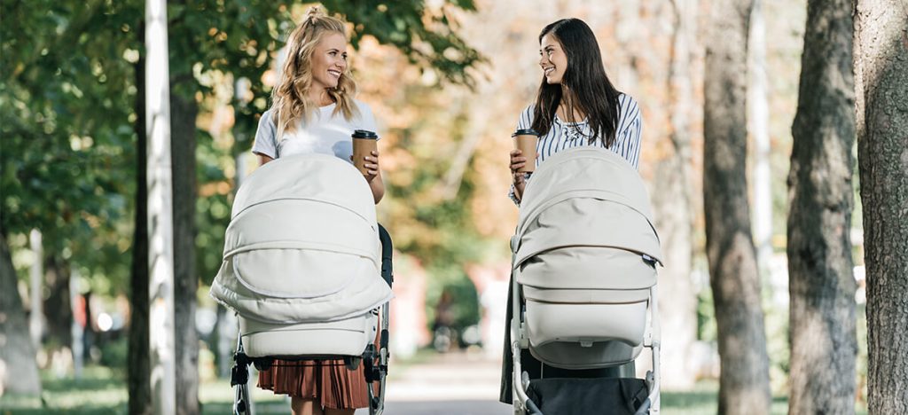 Mothers walking with baby strollers and coffee to go in park and looking at each other