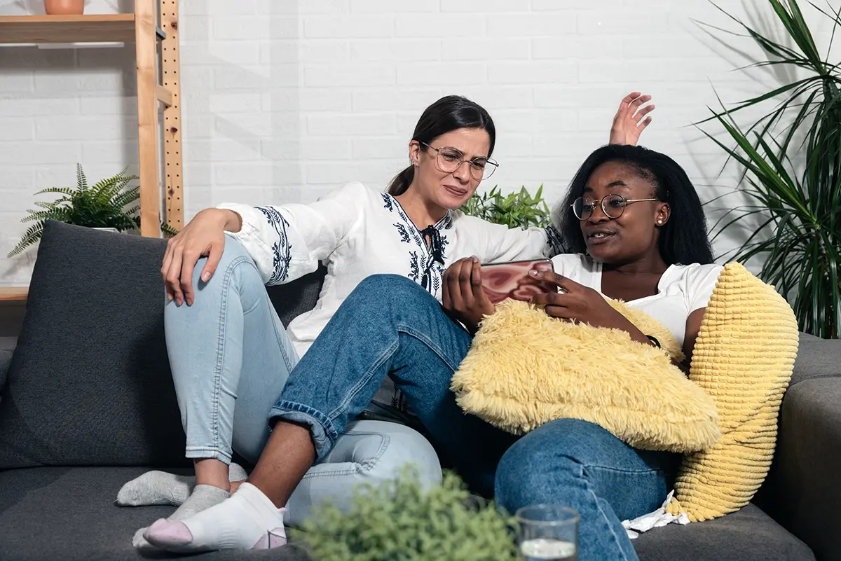 Two young woman sitting on sofa at home and one showing the other video on the smartphone