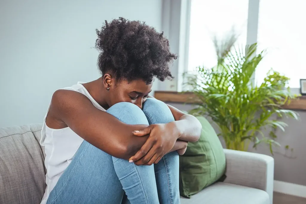 Woman sitting on couch holding her knees to her chest, with her face against her knees looking stressed.