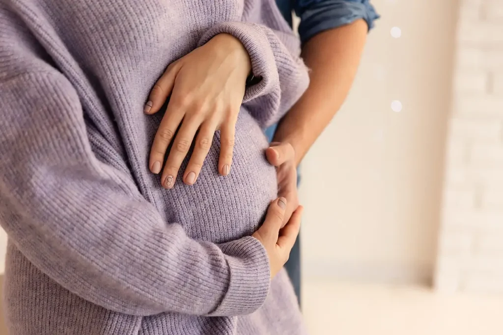 A close-up image of a pregnant woman wearing a soft purple sweater, with her hands gently resting on her baby bump. A second person, likely a partner, stands behind her with their arms wrapped around her, hands also placed supportively on her belly.
