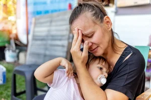 A tired woman sits outside holding a child with a pacifier. She has her hand on her forehead and looks stressed. The child rests in her arms.
