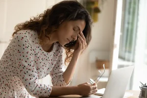 Side view of woman leaning over a table with laptop in front of her grabbing between her eyes.