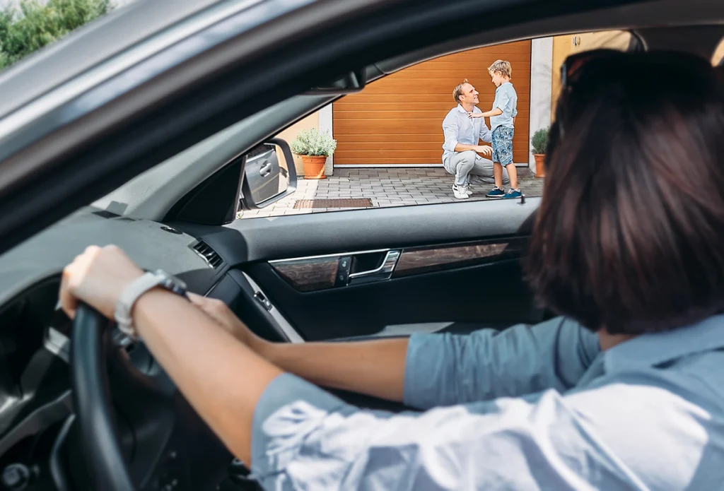 Mother in car looking at their child with their father outside a house,