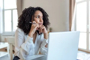 Woman sitting at a desk with a laptop, resting her chin on her hand and holding a pen while looking thoughtfully to the side.