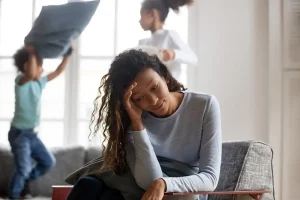 A tired woman sits on a couch with her hand on her forehead, looking stressed, while two children play and have a pillow fight in the background.