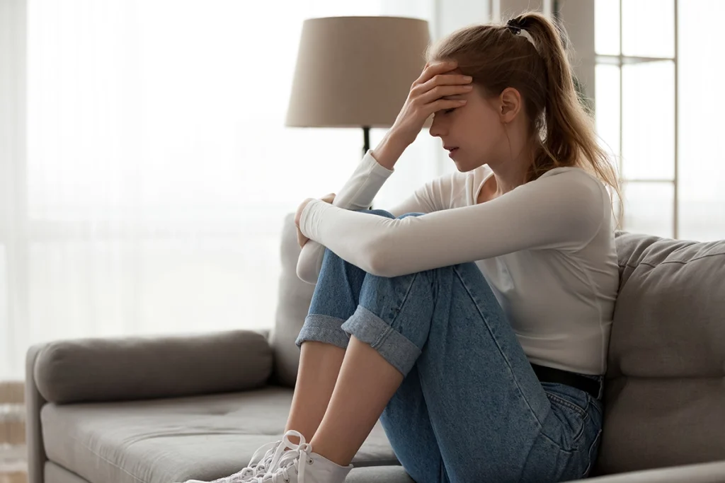 Woman sitting on a couch with her knees pulled close, resting her hand on her forehead and appearing stressed or anxious in a softly lit living room.