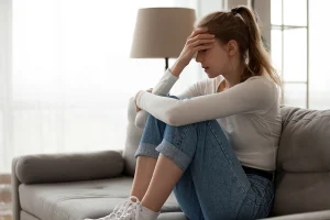 Woman sitting on a couch with her knees pulled close, resting her hand on her forehead and appearing stressed or anxious in a softly lit living room.