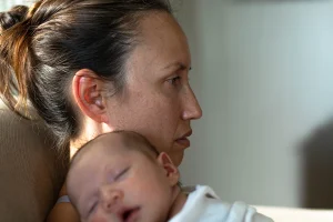 A tired-looking woman sits in profile with her hair tied back, holding a sleeping newborn against her chest. Soft natural light highlights her thoughtful, slightly weary expression.