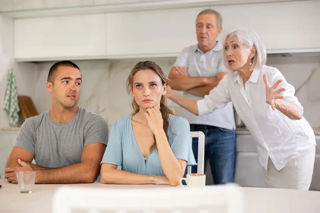 A tense family scene in a kitchen where a young woman sits at a table looking stressed while a man beside her looks concerned, and two older adults argue animatedly in the background.