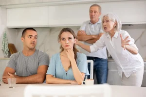 A tense family scene in a kitchen where a young woman sits at a table looking stressed while a man beside her looks concerned, and two older adults argue animatedly in the background.