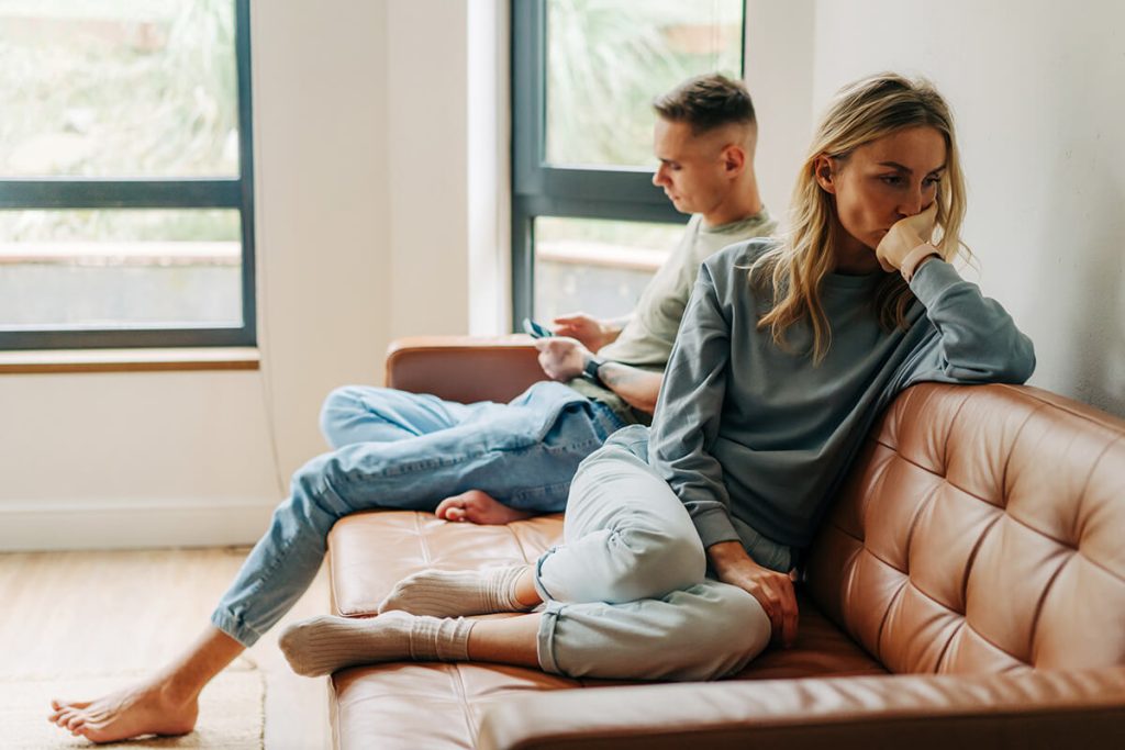 A woman sits on a couch looking upset with her hand against her face, while a man sits beside her turned away, focused on his phone. Both appear distant and tense, suggesting they may be in the middle of an argument or emotional disconnect.