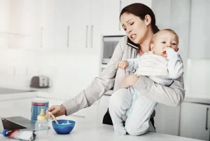 Young mother holding a baby in kitchen while trying to make phone call