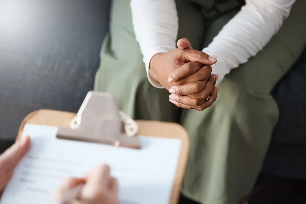 Woman with hands together and therapist writing on clipboard in consultation