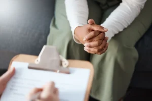 Woman with hands together and therapist writing on clipboard in consultation